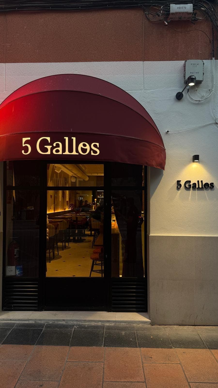 Fachada de 5 Gallos al anochecer: toldo vino con el nombre iluminado y el letrero en la pared