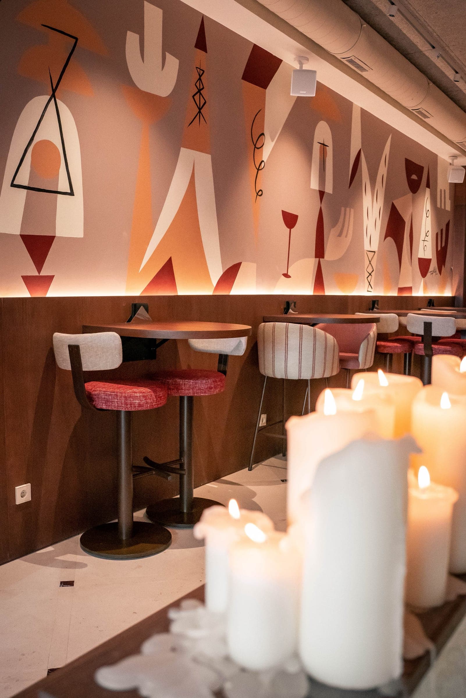 La sala de 5 Gallos: mesas altas de madera y taburetes rojos bajo el mural de Teddy Kelly, con velas en primer plano